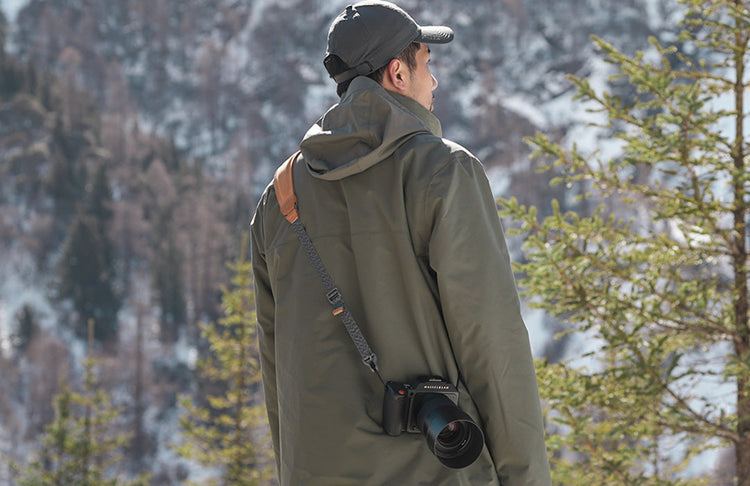 A photographer wearing an outdoor jacket and a ulanzi camera shoulder strap carrying a camera in a snowy forest.