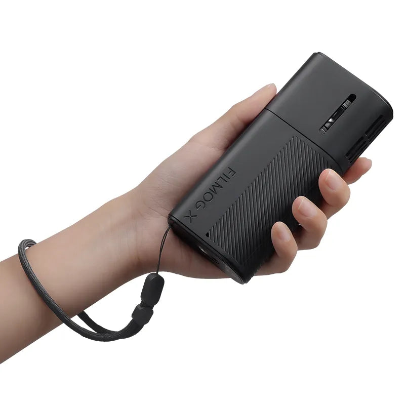 A hand holding a compact FILMOG X portable fog machine with wrist strap.