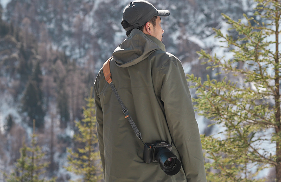 A photographer wearing an outdoor jacket and a ulanzi camera shoulder strap carrying a camera in a snowy forest.