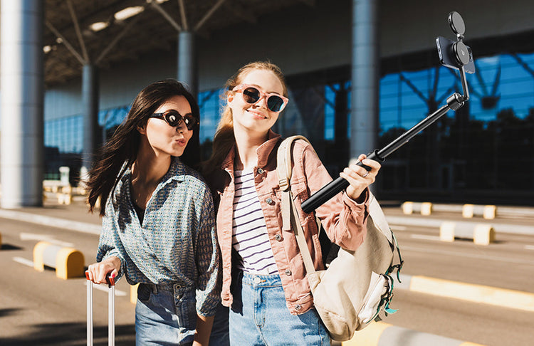 Two young women taking a selfie with a smartphone selfie stick at an airport, wearing sunglasses and carrying travel bags