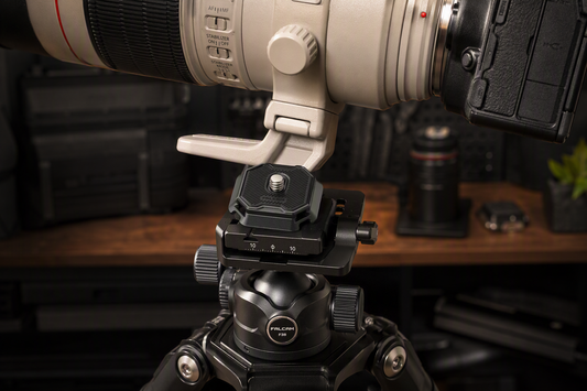 Quick-release plate secured beneath a heavy telephoto lens on a tripod support, with the plate, clamp, and lens foot clearly visible.