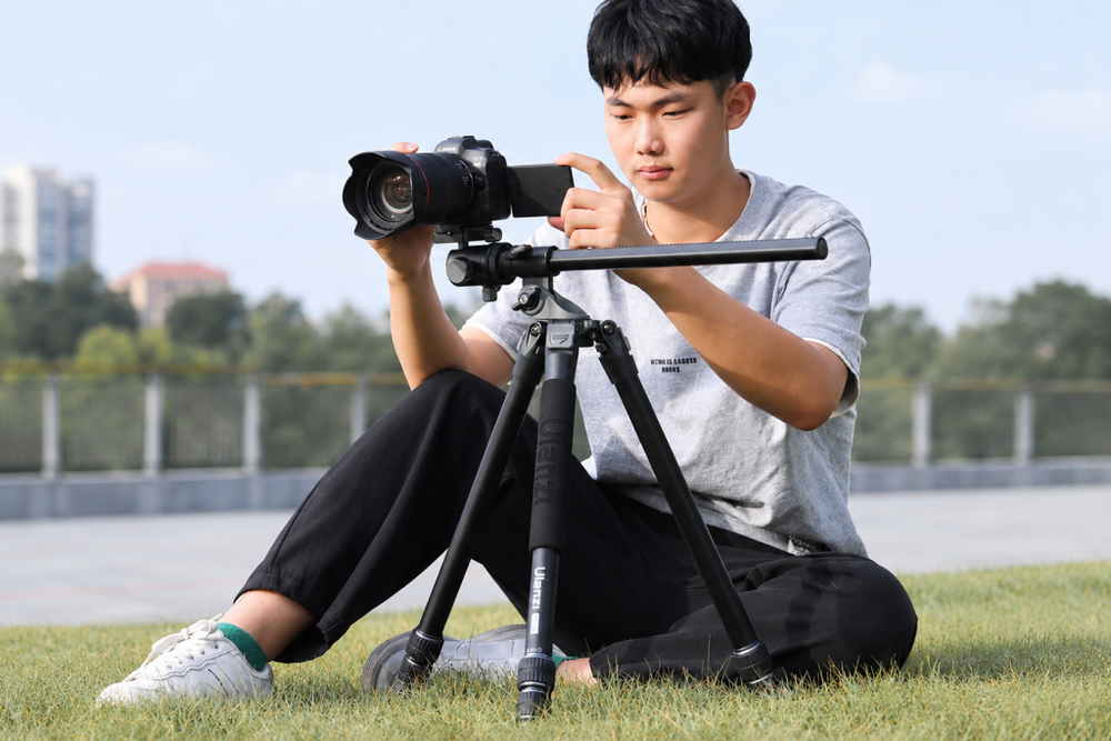 A photographer sitting on the grass while operating a camera mounted on a tripod.