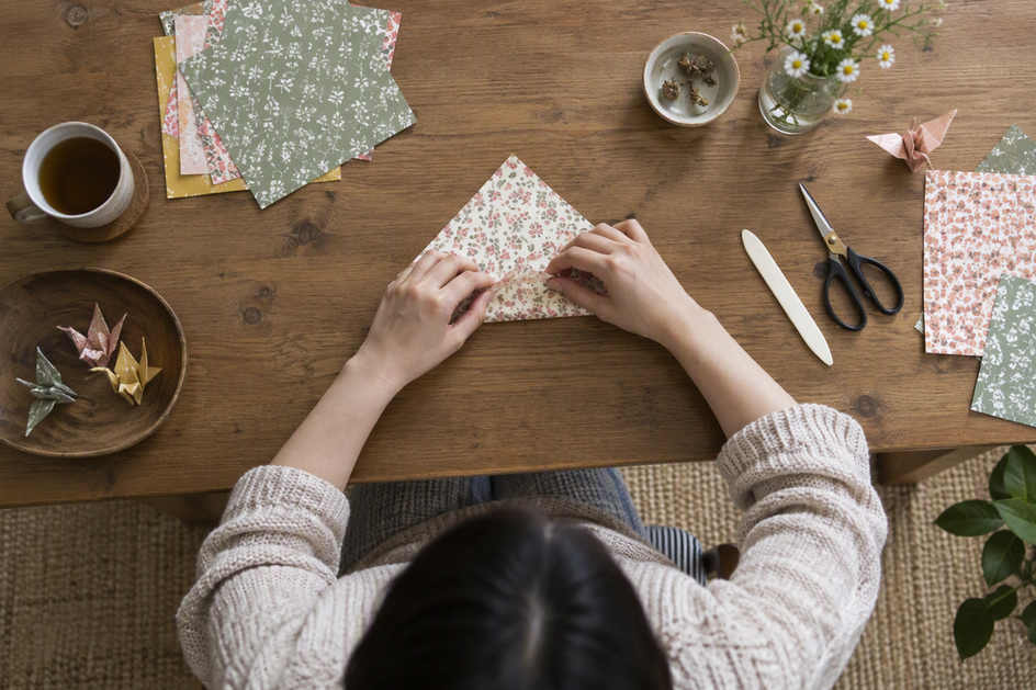 Overhead view of hands folding and arranging patterned paper on a craft table, with the work and hand movements visible.