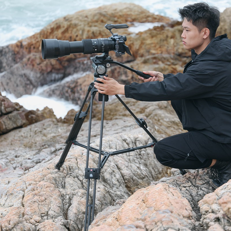User adjusting Ulanzi VideoFast Heavy Duty Tripod Kit on rocky coast with large camera.