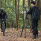 User filming cyclist with Ulanzi VideoFast Heavy Duty Tripod in forest.