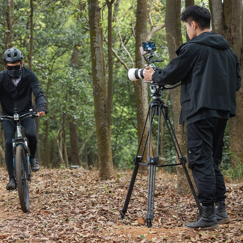 User filming cyclist with Ulanzi VideoFast Heavy Duty Tripod in forest.